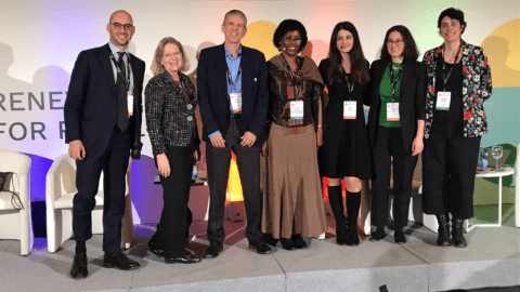 Panellists standing on stage with conference poster as a backdrop